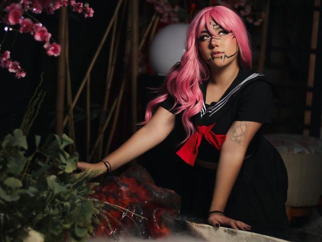 KelseyCoob with pink hair, black sailor dress, red bow, and tattoo on arm, leaning on a floral and red rose-covered surface, looking at camera with headband.