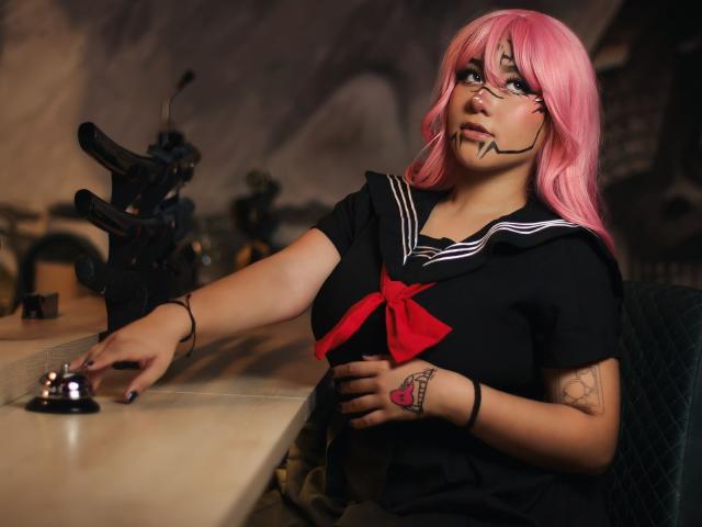 KelseyCoob with pink hair, black sailor top, red bow, and tattoos, touching a joystick, wearing a face mask with black lines, sitting at a desk. Boobs, tits, pussy not visible.
