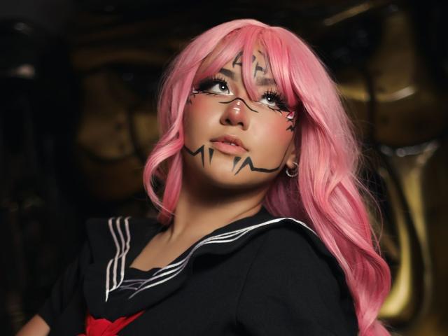 KelseyCoob, avec ses cheveux roses, son uniforme scolaire noir et blanc, ses marques sur le visage en noir, et son rouge à lèvres rose, regarde vers le haut avec une expression légèrement confuse, et porte de petites boucles d'oreilles..