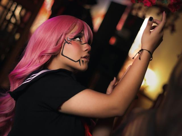 KelseyCoob with pink hair, black eye makeup, and black shirt, touching face with fingers, black nail polish, bracelet on wrist, long wavy hair, side profile, indoors, soft light.