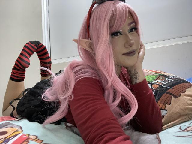 KelseyCoob with pink hair, elf ears, red striped socks, lying on bed, head on hand, looking at camera, black dress, red long sleeves, tattoo on right hand.