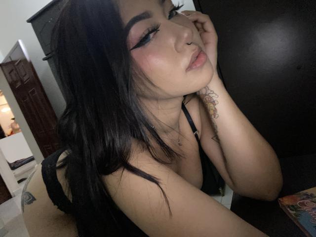 KelseyCoob with black hair, pink eyeshadow, and nose ring, leaning on dark surface, wearing black lingerie, showing tattoo on hand, with soft pink lips and subtle smile.