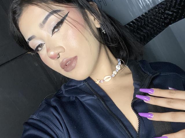 KelseyCoob with dark hair, bold eyeliner, nose ring, glossy lips, purple nails, white shell necklace, black zip-up, and hand on chest, looking at camera.