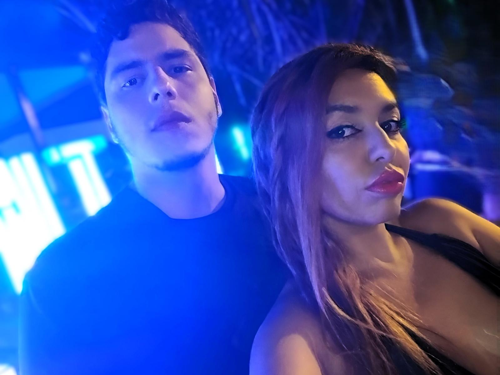 OliverAndCharlotte69 with brown hair, black dress, and pouty lips stands next to OliverAndCharlotte69 with dark hair, black shirt, and neutral expression in blue-lit photo.