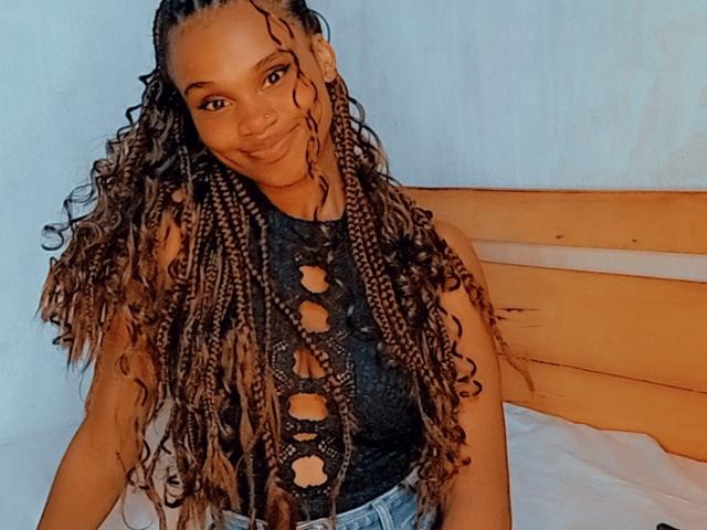 Tiah with long, curly, brown braided hair wearing a black, cut-out sleeve top, smiling at the camera, sitting on a wooden bed with white sheets.