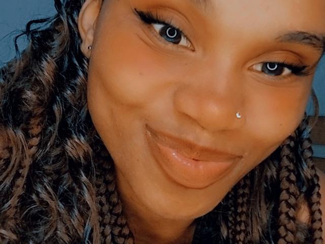 Tiah has brown curly braids, a nose piercing, and a lip piercing with glossy lips, smiling at the camera with blue eyes and dark eyeliner.