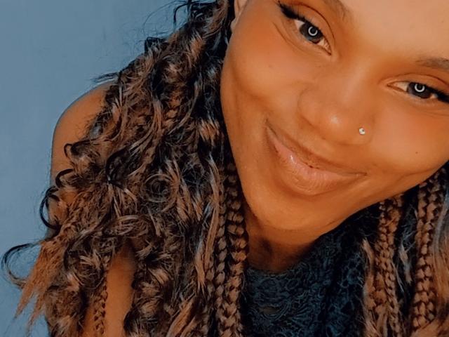 Tiah's smiling face with a small nose piercing, brown eyes, and curly braided hair. Her lips are slightly parted, showing her teeth. She's wearing a dark top.