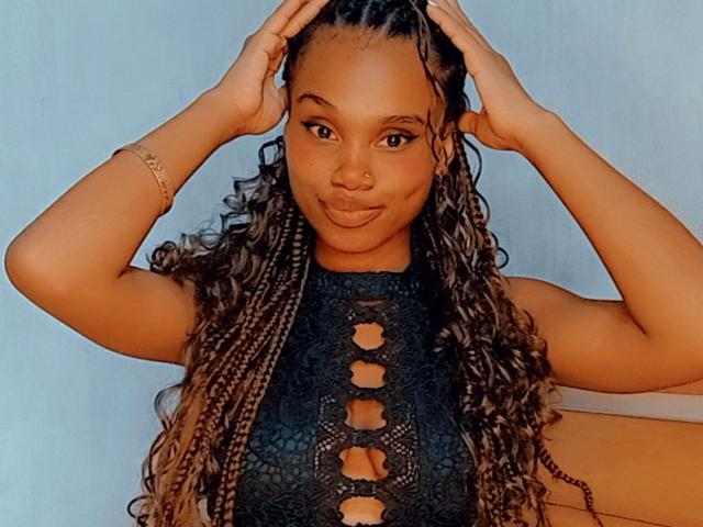Tiah with dark brown skin, long curly braids, wearing a black lace-up top, hands in braids, subtle smile, nose ring, bracelet, and a confident, sexy look.