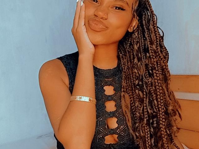 Tiah has dark brown skin, long braided hair, and a cute nose ring. She's wearing a black lace top with cutouts, touching her cheek, and smiling.