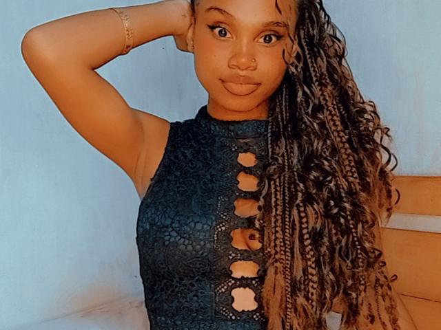 Tiah with brown skin, long curly braids, wearing a black lace-up top, has a cute nose and pouty lips, arm raised, looking at camera, gold bracelet on wrist.