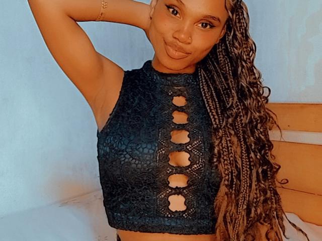 Tiah with tan skin and long braids, wearing a black lace-up crop top, has her arm raised, pouting her lips, and showing her small, round tits.