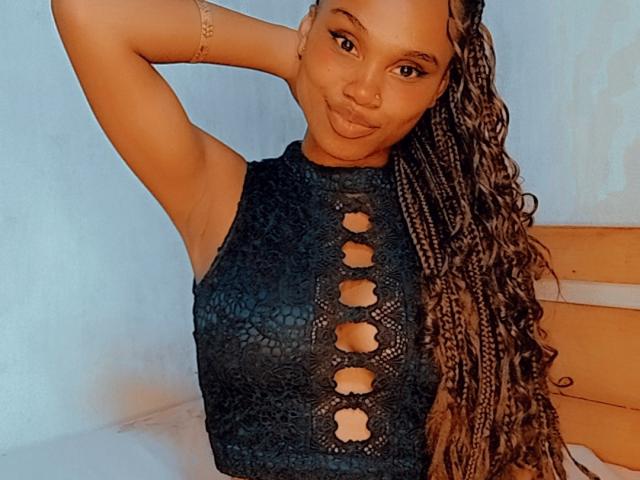 Tiah with brown skin, long braided hair, black lace-up top, and arm raised, smiling at the camera, showing her tits through cut-out top.