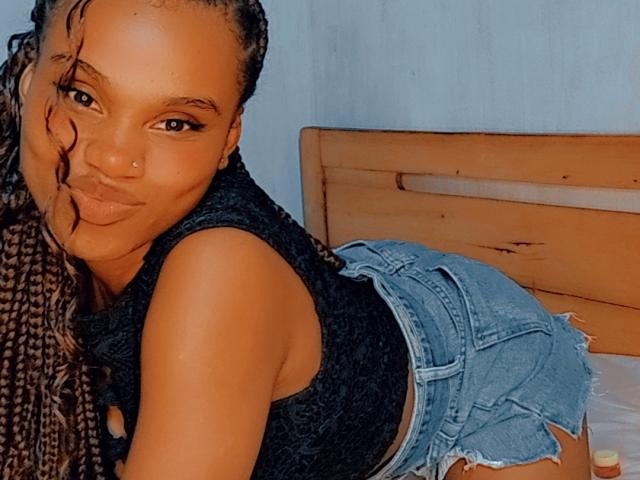 Tiah with her dark brown, braided hair, pouty lips, and nose ring, wearing a black sleeveless top and frayed denim shorts, leaning forward on a wooden bed.