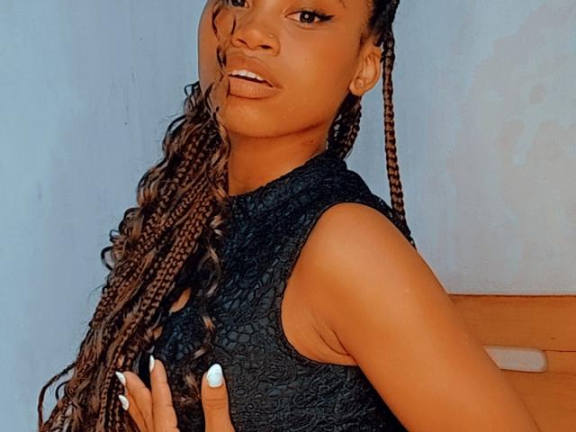 Tiah with brown skin, long braided hair, wearing a black textured sleeveless top, white nails, looking to the side, slight smile, hand on chest, sexy.