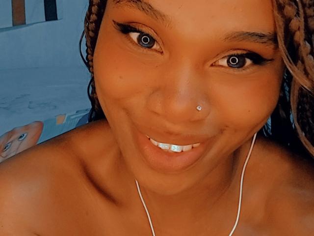Tiah with brown braided hair, nose piercing, and blue eyes, smiling at the camera, showing her white teeth, wearing a silver necklace, and no clothes on.
