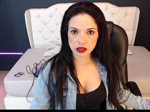 DanielleSandys with long black hair, red lipstick, and a denim jacket over a black top, sitting in a white tufted headboard bed room, looking directly at the camera.