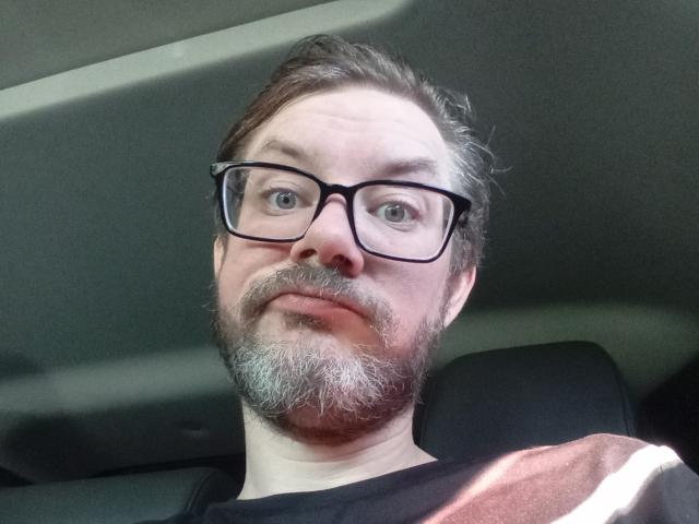 GoreGoblin has a goofy expression, gray beard, and black glasses, with a slight smirk and a red and brown striped shirt. He's sitting in a car.