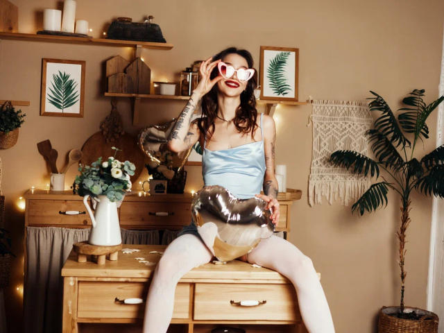 SophieKitty con senos, vestida con una blusa plateada brillante, sosteniendo un espejo, sentada en una mesa de madera, con ropa interior blanca, brazo tatuado, lápiz labial rojo, sonriendo, planta en maceta, pared beige..