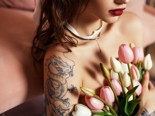 SophieKitty with pink and white tulips, red lipstick, white choker, dragon tattoo on her right arm, holding flowers, upper body shot, brown wavy hair, pink background.
