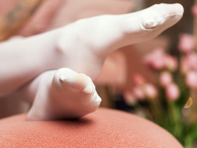 SophieKitty's white, slightly textured foot with a visible toe crease is raised, resting on a pink, textured surface, with blurred pink flowers in the background.