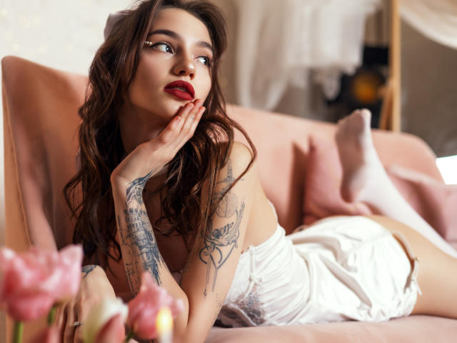 SophieKitty with wavy brown hair, red lipstick, tattoos on arms, lying on pink couch, white dress, hand on cheek, looking up, pink flowers in foreground, soft lighting.