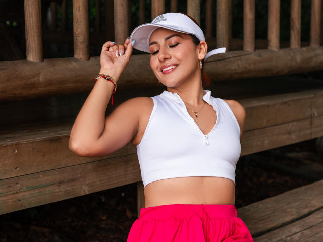 BombSophie with tan skin, brown eyes, and a smile, wearing a white zip-up top, pink shorts, and a white cap, raising her hand, showing a red bracelet.