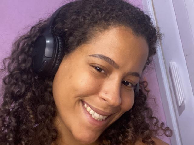 VanessaNataliia with curly brown hair, black headphones, smiling, showing her teeth, and a small nose piercing, against a purple wall. She's looking slightly to the right.