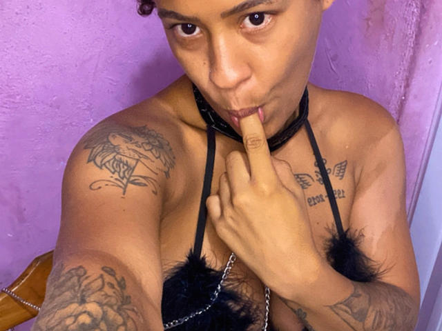 VanessaNataliia with brown skin, short curly hair, black leather collar, black feathered bra, tattoos on shoulder and chest, sucking her finger, visible cleavage, purple wall background.