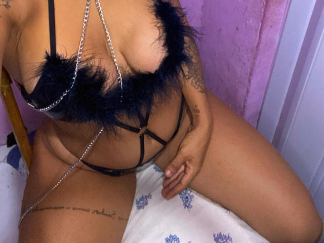 VanessaNataliia with tan skin, black feathered bra, and silver chain, showing her tits and tattoo on thigh, sitting on blue-patterned bed. Her hand rests on her thigh.