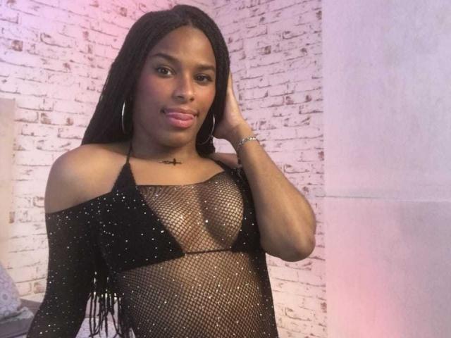 StormyCooper with dark skin, long braids, and a cross necklace, wearing a black mesh top with small sparkles, showing her small boobs, pouty lips, and a relaxed hand on her head.