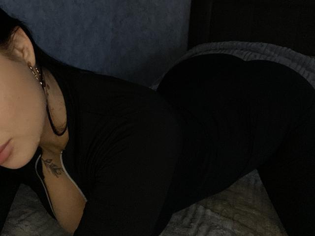 Olieri is lying on a bed, wearing a black outfit, with her butt raised and a tattoo visible on her chest. Her face is partially visible, showing a lip tattoo.