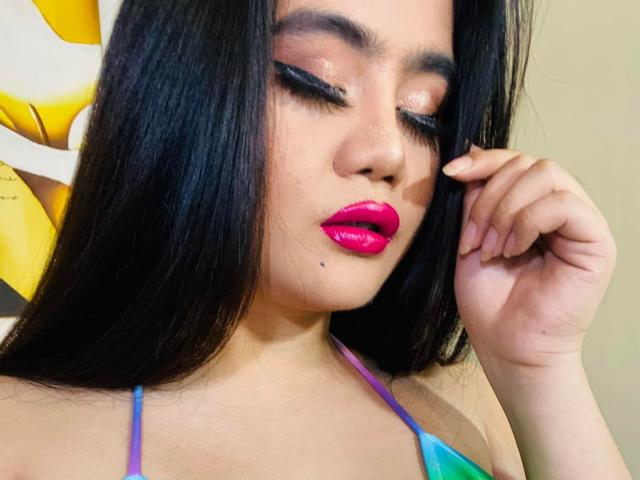 NastyValeria has long black hair, pink glossy lips, and closed eyes with shimmering eyeshadow. She's touching her face with a hand, wearing a colorful halter top.