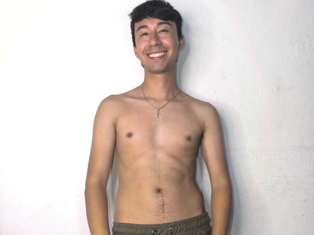 OliverHot69 sin camisa, con un cuerpo delgado y tonificado, pechos pequeños, piel marrón claro, cabello corto y oscuro, sonriendo, usando pantalones verdes y un collar con una cruz, todo frente a una pared blanca..