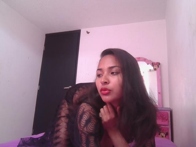 BellaChaudAmour with long black hair, red lipstick, and black lace lingerie, leaning forward, showing her small tits, in a pink-lit bedroom with a mirror and dresser.