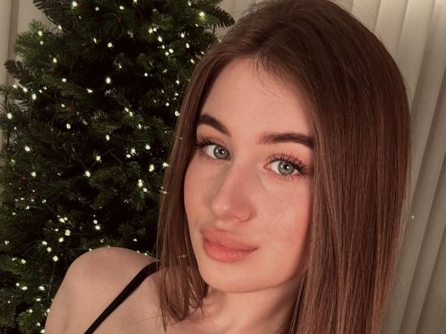 LillyColins heeft bruin haar, groene ogen en roze lippen. Ze draagt een zwarte top en staat voor een kerstboom met kleine witte lampjes..