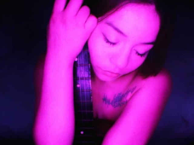 LilithInTheEdenX com pele com tons de rosa, olhos fechados, segurando a alça da guitarra, tatuagens no peito, sem blusa, mão direita perto do rosto, fundo escuro, iluminação suave, tom rosa-roxo..