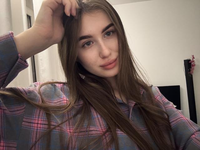 LillyColins met lang bruin haar, die een roze en blauw geruit shirt draagt, kijkt naar de camera met een lichte glimlach, met haar hand in haar haar. Ze heeft blauwe ogen en een lichte huid..