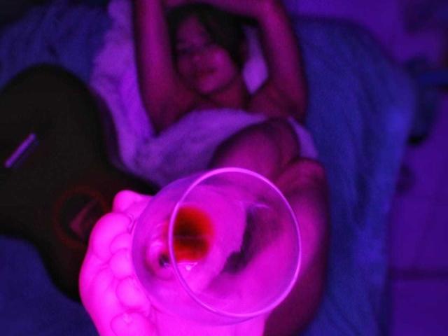 LilithInTheEdenX liegt auf einem Bett, die Arme über dem Kopf, trägt weiße Dessous, im Vordergrund steht ein Glas mit einer roten Flüssigkeit, beleuchtet mit violettem Licht..