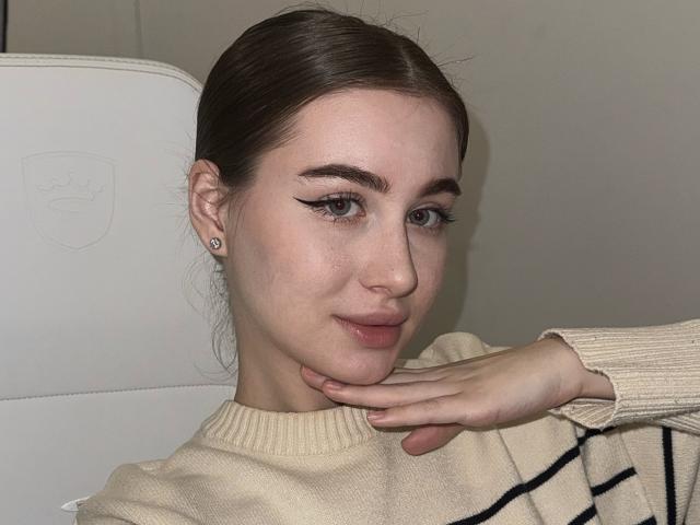 LillyColins met een lichte huid, bruin haar in een lage knot, een cat-eye eyeliner, subtiele roze lippenstift, een beige trui, een hand die haar wang aanraakt, diamanten oorbellen, een zachte glimlach, kijkend naar de camera..