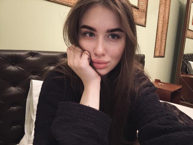 LillyColins with long brown hair, green eyes, and pouty lips, resting her cheek on her hand, wearing a black fuzzy sweater, on a bed with a brown tufted headboard.