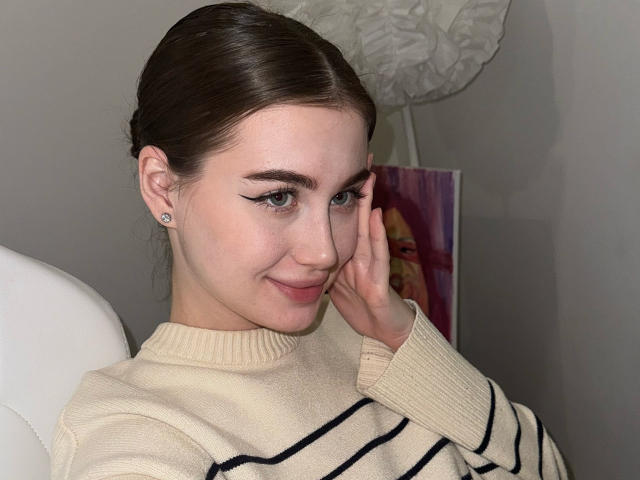 LillyColins con cabello castaño oscuro en una trenza, vistiendo un suéter beige a rayas, tocándose la mejilla, sonriendo, mostrando su delineador de ojos y pequeños aretes, sentada en una silla blanca..