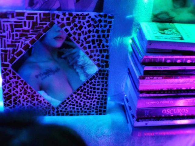 De borsten van LilithInTheEdenX in een spiegel met een mozaïekpatroon, die blauw en paars licht reflecteren, naast een stapel boeken met neonroze en groene accenten..