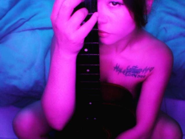 LilithInTheEdenX with purple-pink lighting, holding a guitar, tattoos on chest, sitting naked, looking at camera, guitar strings in front, right hand on neck, left hand on body.