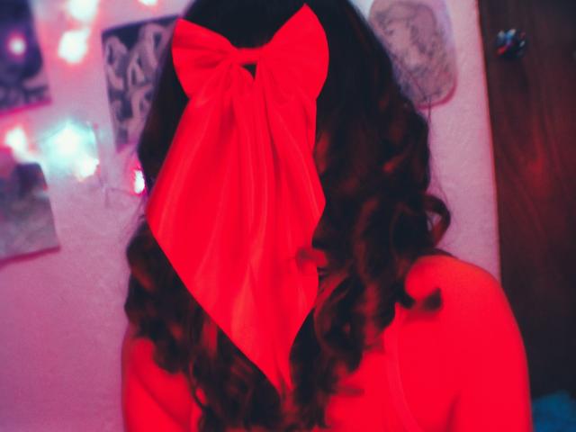 LilithInTheEdenX with curly brown hair, a huge red bow covering her face, and a red-tinted top, showing her bare back and shoulders. Bright, blurry background.