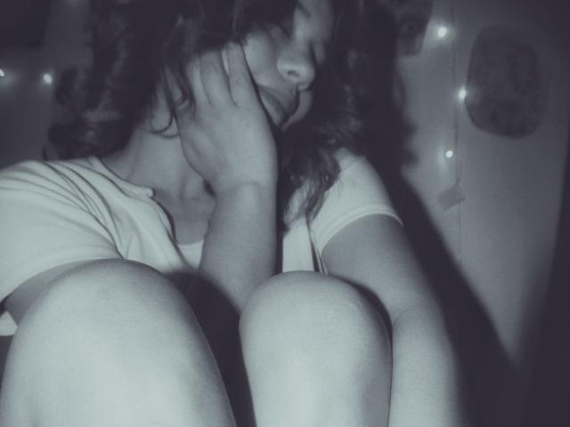 LilithInTheEdenX with curly hair, white shirt, and knees up, smiling softly, hand on cheek, in a grayscale photo. Her tits are small, and she looks cute.