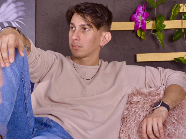 Andrew Carmona tiene el pelo corto y castaño, una camisa beige clara, jeans azules y un reloj de pulsera negro. Está sentado con un cojín rosa y esponjoso, mirando hacia un lado..