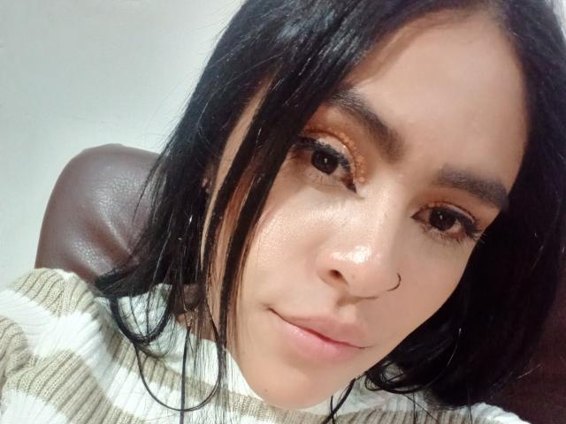 StellaBeckker with black hair, brown eyes, nose ring, and light skin, wearing a striped white and brown sweater, has a slight smile, and a shiny face.