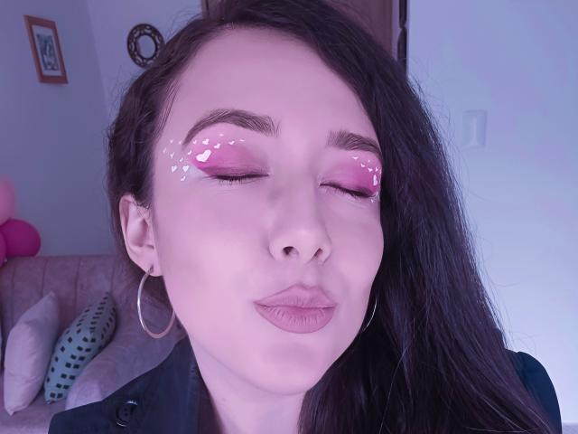 MabyLegrand has pink glittery eyelids, puffy pink lipstick, and is pouting with eyes closed, wearing a black shirt and large hoop earrings, with long dark brown hair.