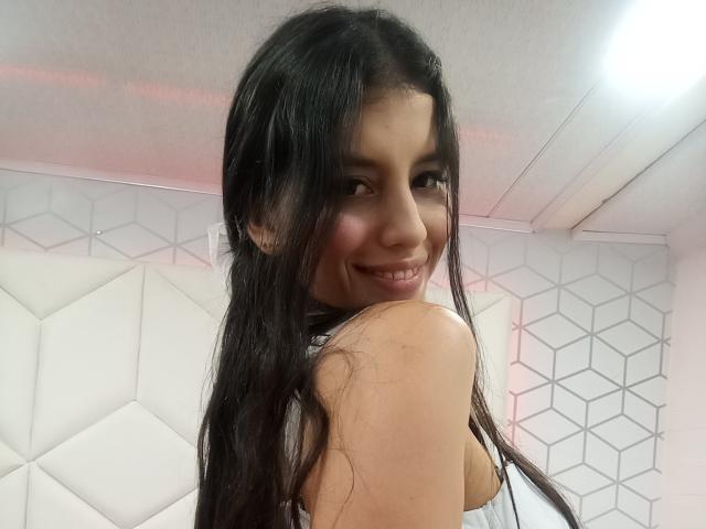 MadissonFontaine with long black hair, smiling at the camera, showing her back and a glimpse of her tits, wearing a white top, in a brightly lit room.