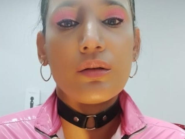 PaulaCamis has dark skin, pink eyeshadow, and glossy lips. She's wearing a pink shiny jacket, black choker with metal ring, and hoop earrings. Her expression is neutral.
